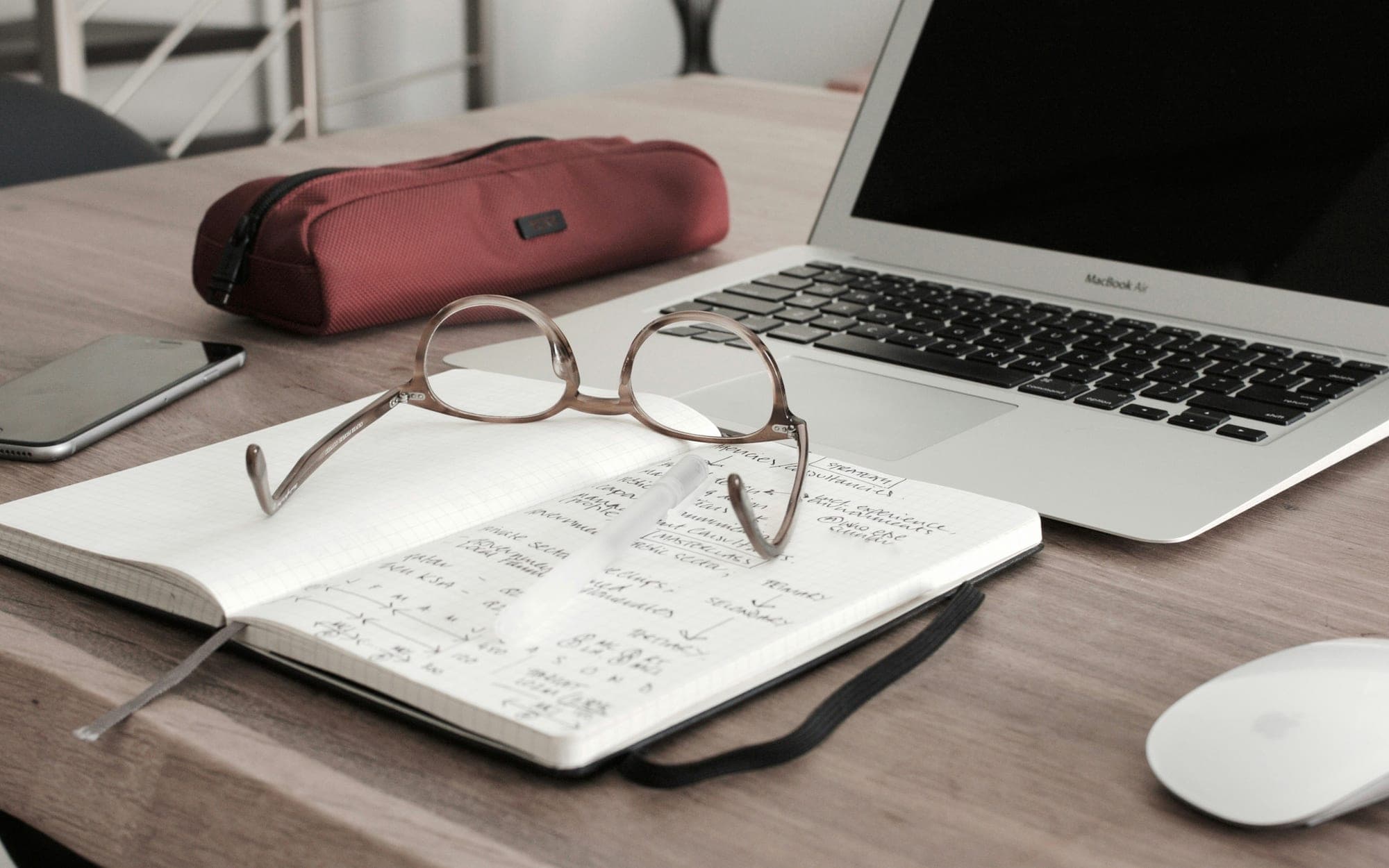 Reading glasses resting on a notebook of handwritten notes beside a laptop